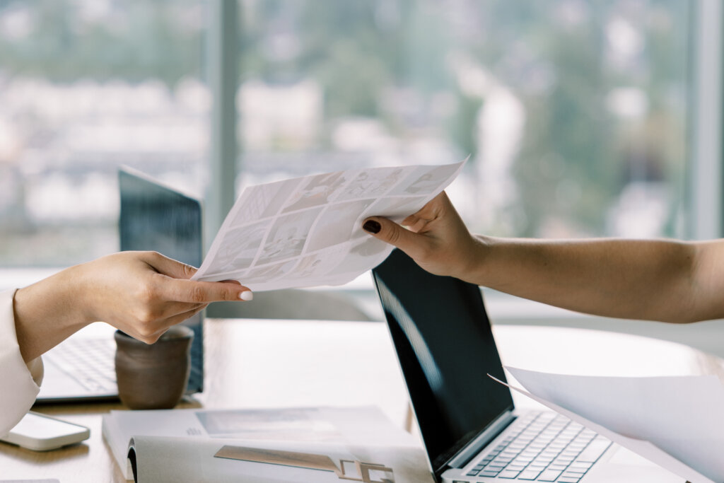 What Are Backlinks? (And How To Grow Them Authentically) Two Business Owners Passing A Document Across A Desk With A Laptop, Collaborating To Earn Backlinks