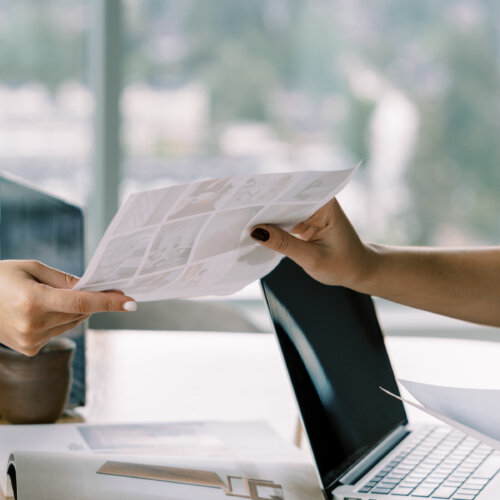 Two business owners passing a document across a desk with a laptop, collaborating to earn backlinks